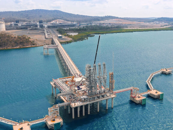 Aerial view of an offshore LNG terminal jetty and loading platform connected to shore, showing pipelines, storage tanks, and service walkways over blue water