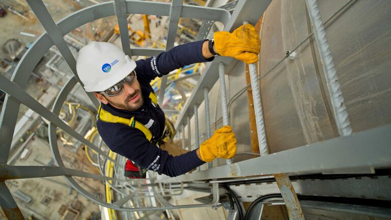 Un homme portant un casque de sécurité avec le logo de T.EN grimpe sur une échelle d'un site industriel