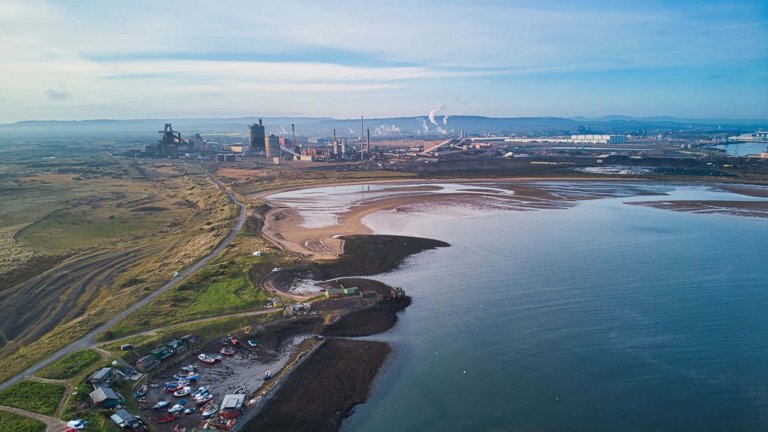 Vue aérienne d'un paysage industriel avec des usines près d'un plan d'eau calme, une plage de sable et des collines verdoyantes au premier plan