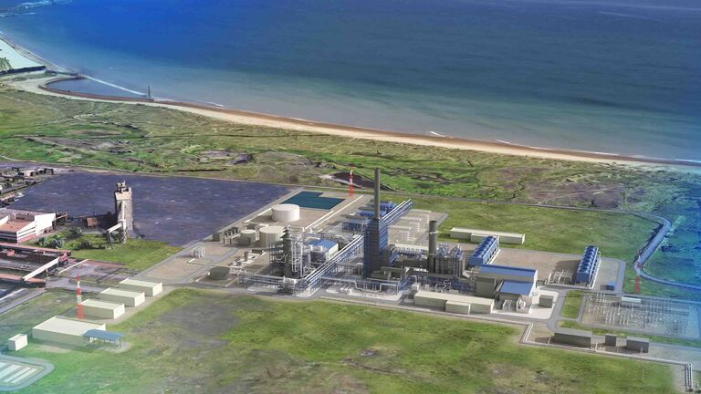 Aerial view of a large industrial facility located near the coast, with various buildings and a sandy beach in the background