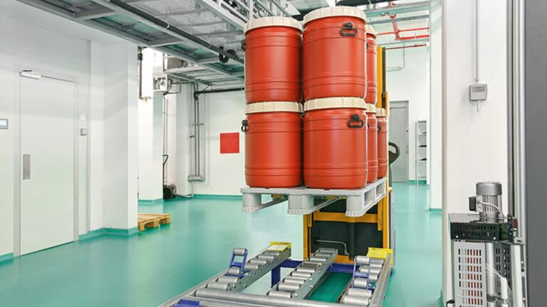 A rack system lifts large orange barrels in a clean industrial space with green flooring and bright lighting