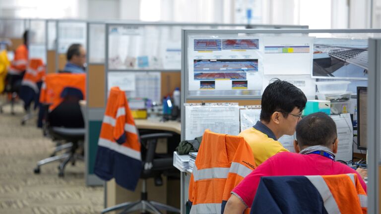 Two workers in safety vests collaborate at desks in a busy office environment, focused on their computer screens