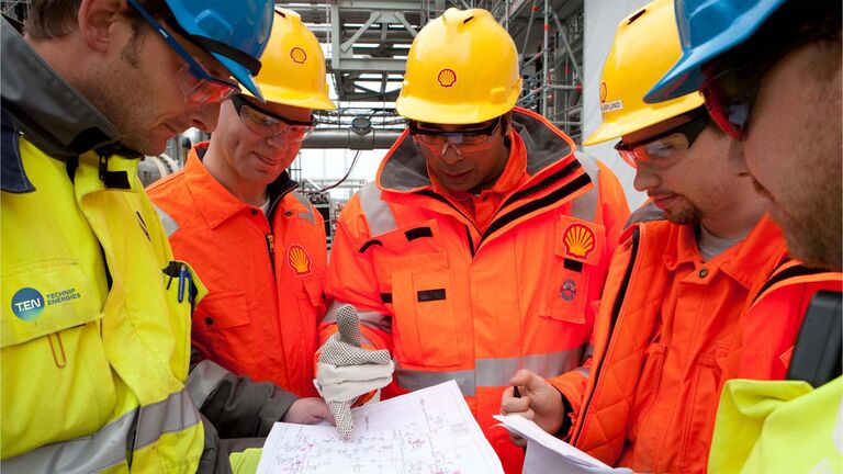 A group of workers in bright orange safety jackets and yellow helmets discussing plans on a construction site