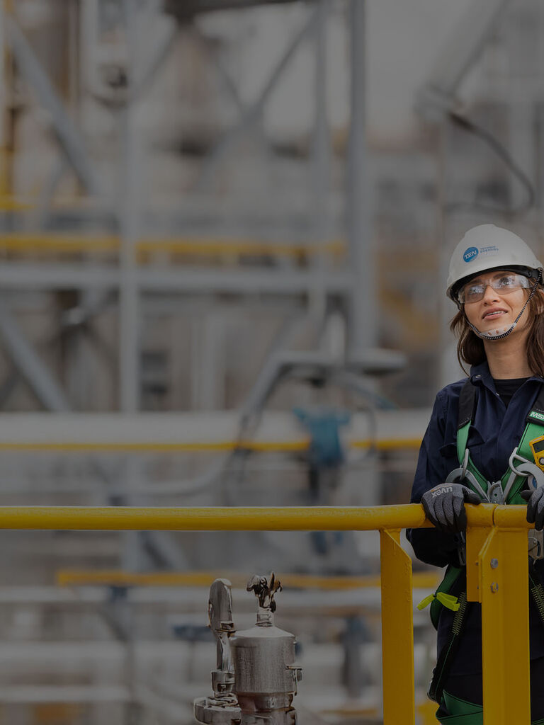 Three workers in safety gear stand on a platform, smiling, with industrial equipment visible in the background