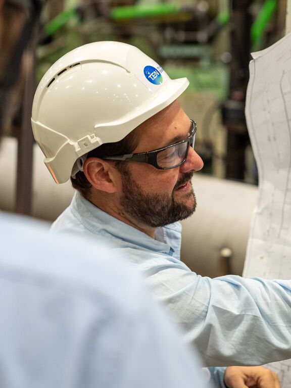 Engineer in hard hat and safety glasses reviewing technical blueprints with a colleague inside an industrial energy plant