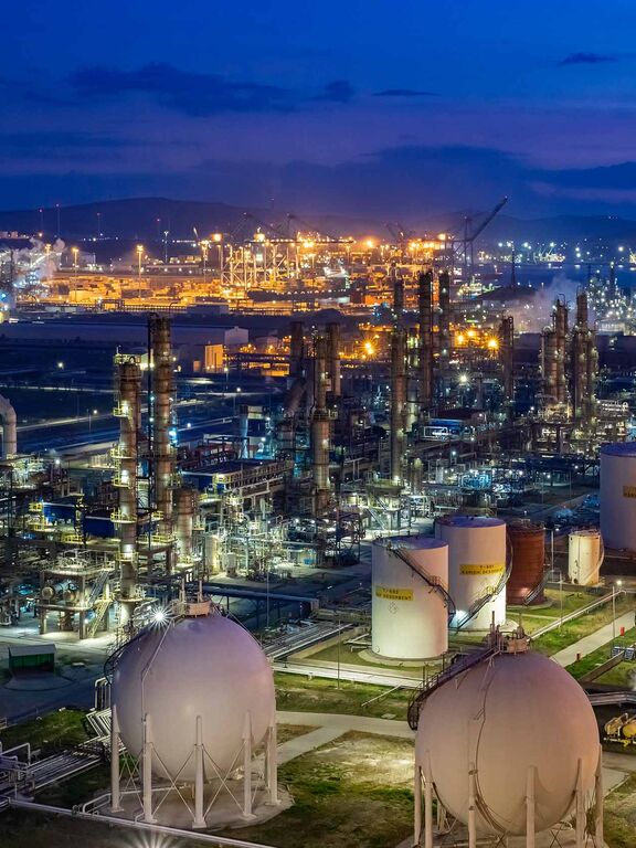 Illuminated oil refinery and petrochemical complex at dusk, with processing towers, storage tanks, and industrial infrastructure.