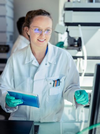 A scientist in a white lab coat smiling, holding a notebook while looking at a screen