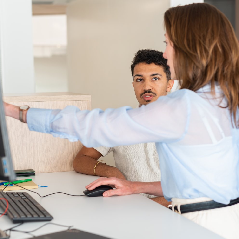 Woman in blue shirt pointing to computer screen while a young man with a moustache looks at her