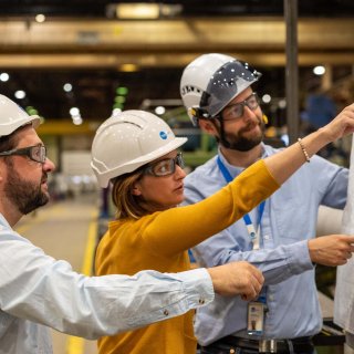 Trois professionnels portant un casque de sécurité discutent de plans dans une usine, mettant en avant le travail d'équipe en ingénierie