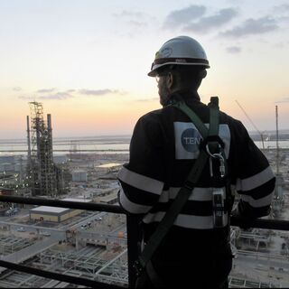 A worker in a safety helmet and harness stands on a platform overlooking an industrial facility at sunset, with various structures and equipment visible in the background