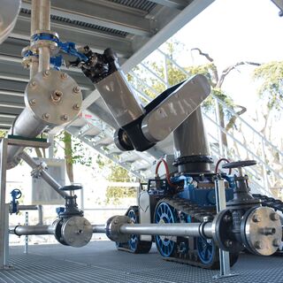 A robotic system with articulated arms and wheels, positioned on a metal platform, surrounded by pipes and machinery