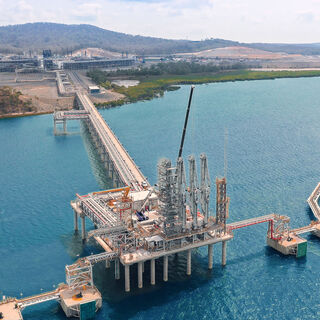 Aerial view of an offshore LNG terminal jetty and loading platform connected to shore, showing pipelines, storage tanks, and service walkways over blue water