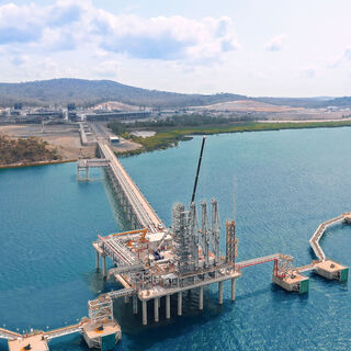 Aerial view of an offshore LNG terminal jetty and loading platform connected to shore, showing pipelines, storage tanks, and service walkways over blue water