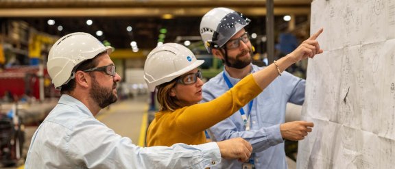 Three professionals wearing safety helmets discussing plans in a factory, emphasizing teamwork in engineering