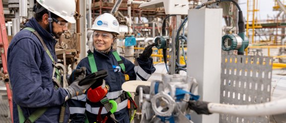 Two workers (a woman and a man) wearing a helmet with the Technip Energies logo on a construction site, taking notes about a piece of equipment in front of them