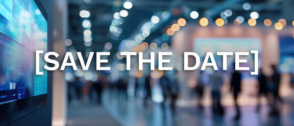 A blurred view of a large indoor event space filled with people and warm overhead lights, creating a soft bokeh effect. On the left side of the image, a digital display screen is partially visible. Centered across the image is the bold white text “[SAVE THE DATE]”.