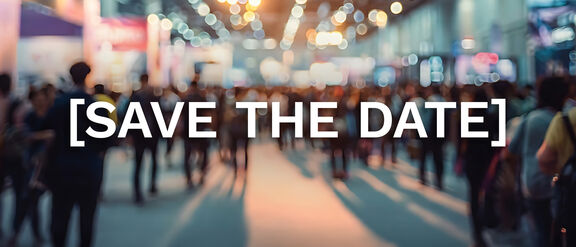 Blurred view of a busy event hall filled with people walking through an exhibition space, illuminated by bright overhead lights, with the words ‘SAVE THE DATE’ displayed prominently in the center.