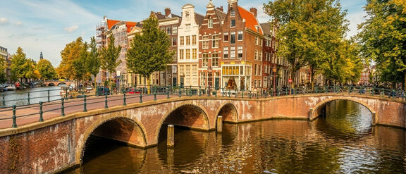 Arched brick bridge over a canal in Amsterdam with traditional narrow houses and trees along the water.