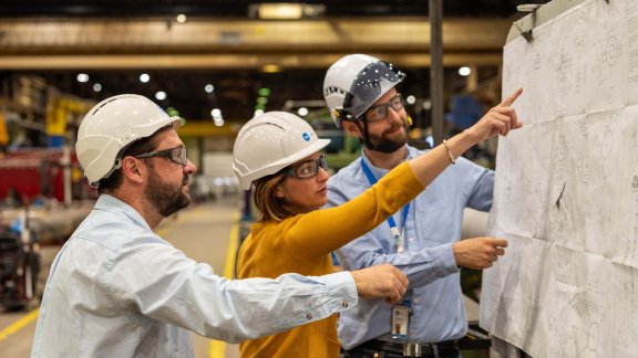 Three professionals wearing safety helmets discussing plans in a factory, emphasizing teamwork in engineering