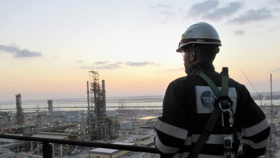A worker in a safety helmet and harness stands on a platform overlooking an industrial facility at sunset, with various structures and equipment visible in the background