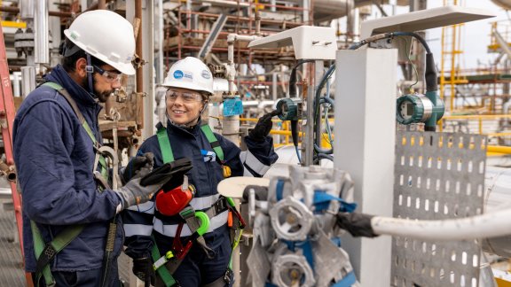 Two workers (a woman and a man) wearing a helmet with the Technip Energies logo on a construction site, taking notes about a piece of equipment in front of them
