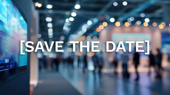 A blurred view of a large indoor event space filled with people and warm overhead lights, creating a soft bokeh effect. On the left side of the image, a digital display screen is partially visible. Centered across the image is the bold white text “[SAVE THE DATE]”.