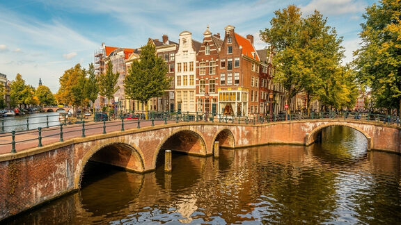 Arched brick bridge over a canal in Amsterdam with traditional narrow houses and trees along the water.
