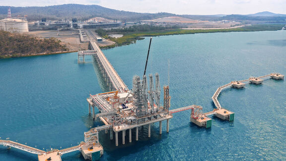 Aerial view of an offshore LNG terminal jetty and loading platform connected to shore, showing pipelines, storage tanks, and service walkways over blue water